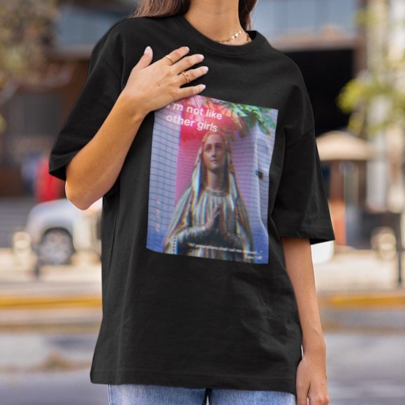 May include: Black t-shirt featuring a graphic print of a woman in a blue robe with the text "I'm not like other girls" in pink and white.