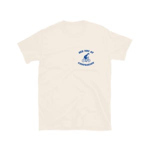 May include: Cream-colored t-shirt with a blue graphic of a person riding a bicycle and the text "SEE YOU AT CONFESSION". The short-sleeved shirt is made of a soft material.