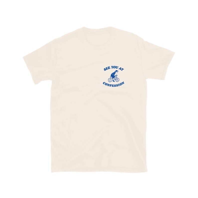 May include: Cream-colored t-shirt with a blue graphic of a person riding a bicycle and the text "SEE YOU AT CONFESSION". The short-sleeved shirt is made of a soft material.