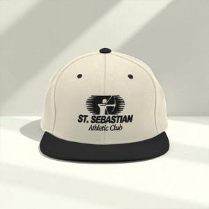 May include: A cream-colored flat-brimmed baseball cap with a black brim and top button. The cap features a black logo with the text "ST. SEBASTIAN Athletic Club" and an archer with a bow and arrow.