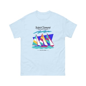 May include: Light blue t-shirt featuring a graphic of three sailboats with colorful sails against a blue and purple background. The text "Saint Clement - Patron Saint of Mariners - Ora Pro Nobis" is printed above and below the sailboat graphic.