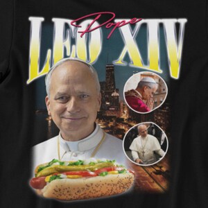 K&ouml;nnte beinhalten: Schwarzes T-Shirt mit einer Grafik eines Mannes, eines Hotdogs und den Worten "LEO XIV" in Gelb und "Pope" in Rosa. Zwei kleinere Bilder des Mannes sind ebenfalls enthalten. Der Hotdog hat Senf, Relish und Tomaten.