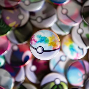 Puede incluir: Primer plano de un pin de Poké Ball colorido con un diseño de acuarela. El pin está rodeado de otros pines de Poké Ball con diferentes diseños.