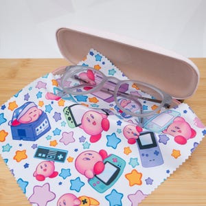Puede incluir: Un par de gafas con montura transparente descansa sobre un paño de microfibra con un colorido estampado de personajes rosas de Kirby, consolas de videojuegos y estrellas. Un estuche para gafas rosa claro está abierto en el fondo.