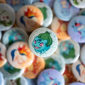 Peut inclure: Une collection de boutons ronds colorés représentant des personnages de dessins animés de la franchise Pokémon. Les boutons sont disposés en pile, avec un bouton vert et bleu représentant une image de dessin animé de Bulbizarre au centre.