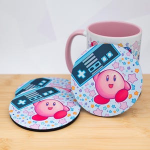 Peut inclure: Ensemble de quatre dessous de verre ronds avec un personnage de dessin animé rose et un motif de manette. Les dessous de verre ont un fond bleu clair avec des étoiles colorées. Une tasse assortie est également visible.