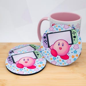 May include: Set of three round coasters featuring a pink Kirby character holding a video game console. The coasters have a light blue background with star shapes. A matching mug with a pink handle and rim is also visible.