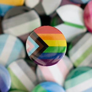 Può includere: Una collezione di spille colorate con vari design di bandiere arcobaleno, tra cui una bandiera arcobaleno, una bandiera transgender e una bandiera bisessuale.