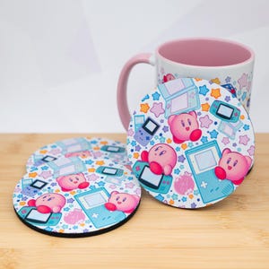 May include: Set of four round coasters featuring a colorful design with pink cartoon characters, blue handheld game consoles, and stars. A matching pink and white mug is in the background. The coasters are on a wooden surface.