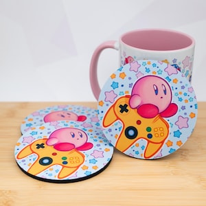 May include: A set of three round coasters and a mug featuring a pink cartoon character with a yellow video game controller design. The coasters and mug have a light blue background with star shapes.