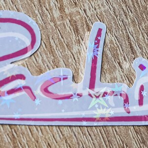 K&ouml;nnte beinhalten: Ein wei&szlig;er Aufkleber mit rosa Umrandung und holografischem Finish. Der Aufkleber zeigt den Namen "Rachel" in einer kursiv geschriebenen Schrift mit Sternen, die um den Text verstreut sind.