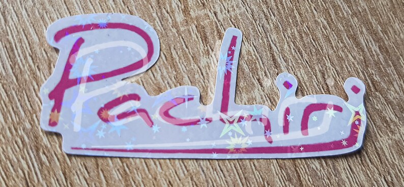 K&ouml;nnte beinhalten: Ein wei&szlig;er Aufkleber mit rosa Umrandung und holografischem Finish. Der Aufkleber zeigt den Namen "Rachel" in einer kursiv geschriebenen Schrift mit Sternen, die um den Text verstreut sind.