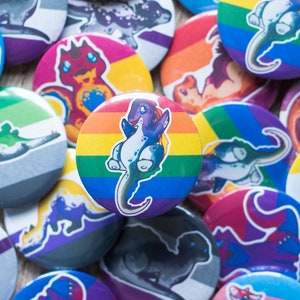 Può includere: Una collezione di bottoni rotondi e colorati con illustrazioni di dinosauri. I bottoni presentano vari disegni di bandiere del pride, tra cui bandiere arcobaleno, bisessuali e asessuali. I dinosauri sono raffigurati in uno stile cartone animato con una varietà di colori.