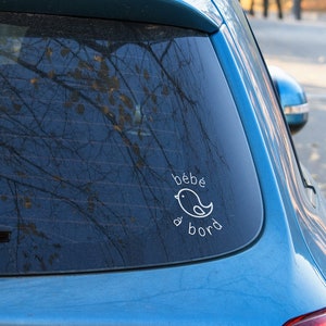 Peut inclure: Une voiture bleue avec un autocollant blanc sur la vitre arrière qui dit "bébé à bord" avec une petite illustration d'oiseau.