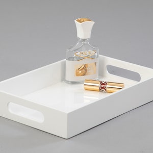 May include: White rectangular tray with two handles on the sides. The tray has a bottle of perfume and a gold lipstick on it.