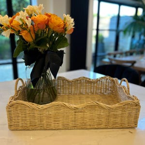 May include: A woven wicker tray with a scalloped edge and two handles. The tray is light brown and is sitting on a white surface. A glass vase with a black ribbon is holding a bouquet of orange and white flowers.