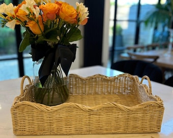 May include: A woven wicker tray with a scalloped edge and two handles. The tray is light brown and is sitting on a white surface. A glass vase with a black ribbon is holding a bouquet of orange and white flowers.