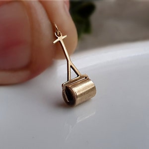 May include: A gold-toned charm in the shape of a miniature roller, with a handle and wheels. The charm is a detailed representation of a small, handheld roller.