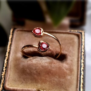 Peut inclure: Une bague ouverte en or dotée de deux sertissages en forme de cœur, chacun contenant une pierre précieuse rouge vive à facettes. L'anneau est lisse et poli, avec les deux motifs en cœur positionnés aux extrémités ouvertes de la structure.
