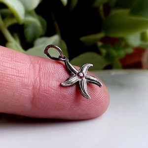May include: Silver starfish charm with a red gemstone in the center. The charm is on a finger.