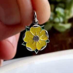 May include: A silver charm in the shape of a yellow flower with green accents. The flower has five petals and a small center.