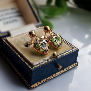 May include: A pair of gold-colored heart-shaped earrings with green accents, displayed in a vintage blue jewelry box with gold trim. The earrings have a stud design and are set against a light beige interior.
