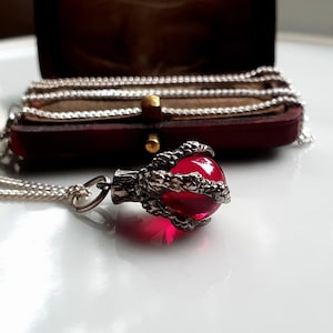 Puede incluir: Un collar de plata con un colgante. El colgante presenta una gema esférica roja brillante sujeta por un engaste en forma de garra. La cadena es fina y el colgante es el punto focal. En el fondo, una caja de joyería de terciopelo rojo.