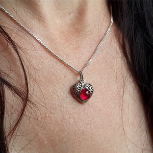 May include: A silver chain necklace with a heart-shaped pendant featuring a red gemstone.