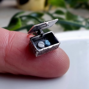 May include: A small, open silver chest with a hinged lid, containing two small, round pills, one gray and one blue. The chest is held between fingers, showcasing intricate detailing and a miniature design, perfect for a unique jewelry piece or charm.