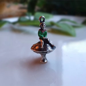 May include: A silver charm depicting a seated figure with a green jacket and red details, sitting on a mushroom. The charm has a loop for attaching to a chain. The mushroom has a detailed, textured surface. The charm is likely for a necklace or bracelet.