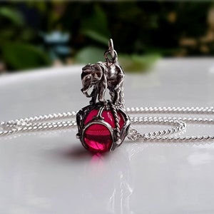 Op de afbeelding: Een zilverkleurige hangerketting met een klein olifantfiguurtje dat op een doorschijnende, bolvormige, felroze kraal staat. De kraal is gevat in een decoratieve zilverkleurige metalen kooistructuur en de hanger hangt aan een zilverkleurige ketting.
