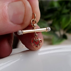 May include: A gold-colored pendant with a horizontal bar and a dangling heart-shaped charm. The heart is red with a gold filigree design. The ends of the bar have a diamond-like texture.
