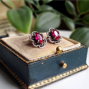 May include: A pair of vintage silver stud earrings, each featuring an oval, deep red gemstone. The earrings have an ornate, openwork design. They are presented in a blue jewellery box with gold trim and a beige velvet interior.