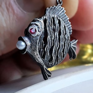 May include: A silver fish pendant featuring a pearl and a red gemstone. The fish is detailed with textured scales and a fin.