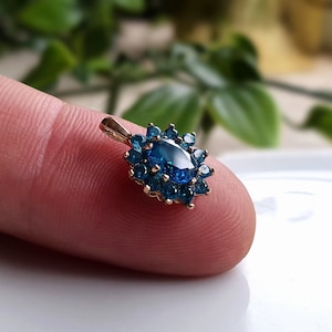 May include: A gold pendant featuring a central oval-cut blue gemstone surrounded by a halo of smaller round-cut blue stones. The pendant has a small metal bail at the top for attachment to a chain, showcasing a vibrant blue color palette.