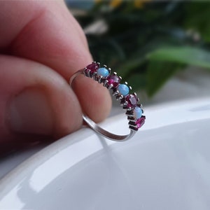 May include: A silver ring with alternating red and light blue gemstones. The ring features a row of small, round gemstones set closely together. The gemstones are a deep red and a pale blue, creating a contrasting colour pattern.