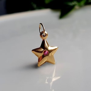 Pode incluir: Um berloque de estrela dourada com uma pequena pedra preciosa vermelha redonda no centro. O berloque tem uma argola na parte superior para pendurar, provavelmente um pingente ou um componente de brinco. A estrela é uma forma clássica, adequada para joias.