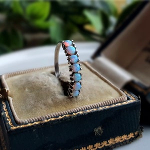 May include: A silver ring set with six oval opals, each displaying blue and orange hues. The ring is presented in a vintage dark blue jewellery box with gold edging and a beige interior.