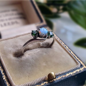 Op de afbeelding: Zilveren ring met een centrale opaal en twee groene edelstenen. De ring heeft een vintage ontwerp en wordt gepresenteerd in een blauwe sieradendoos met een gouden sluiting.