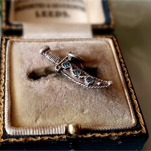 Può includere: Un ciondolo a forma di pugnale in argento con pietre preziose blu incastonate nella lama, esposto in una scatola foderata in velluto.