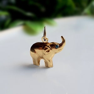 Può includere: Un ciondolo a forma di elefante color oro con una piccola pietra preziosa blu rotonda incastonata come occhio. Il ciondolo presenta una finitura metallica lucida e include un anellino circolare sulla parte superiore per l'aggancio.