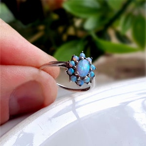 May include: A silver ring featuring a central oval-shaped iridescent opal surrounded by a cluster of ten smaller round opals in a floral arrangement. The metal band is thin and polished, showcasing the blue and white flashes of the gemstones.