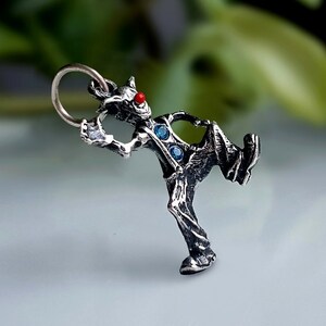 Peut inclure: Pendentif argenté représentant une figurine stylisée avec un nez en gemme rouge et deux gemmes bleues sur la poitrine. La figurine est dans une pose dynamique, avec une boucle en haut pour une chaîne. Finition texturée.
