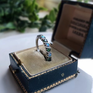 May include: A silver ring featuring an alternating pattern of round, iridescent opals and deep green gemstones set within a curved, wave-like metal band. The ring is displayed upright in a dark blue, velvet-lined jewelry box with gold trim.