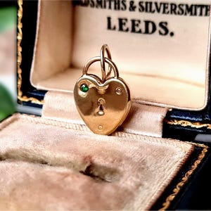 May include: A gold heart-shaped padlock pendant featuring a small green gemstone set in the upper left corner and a keyhole design in the centre. The pendant is displayed inside a black jewellery box with the text GOLDSMITHS & SILVERSMITHS LEEDS.