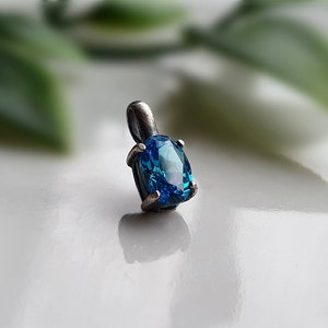 May include: A silver pendant featuring a vibrant, oval-cut blue gemstone. The gemstone is set in a simple, four-prong setting, and the pendant has a small loop for a chain. The background is blurred, with hints of green and white.