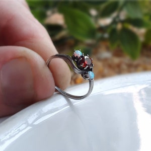 May include: A silver ring featuring a red gemstone and two white opals. The ring has a delicate, curved design.