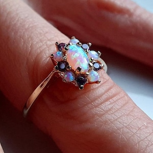 May include: A gold ring with an oval opal centre stone, encircled by smaller, round, purple and opal-coloured gemstones. The ring's design resembles a flower or starburst, with the gemstones clustered together. The ring is shown on a finger.