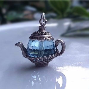Op de afbeelding: Een miniatuur theepotbedel met een facetgeslepen, lichtblauwe kristallen body. De theepot heeft sierlijke details van zilverkleurig metaal op het deksel, handvat en basis. De bedel heeft een lus aan de bovenkant.