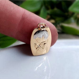 May include: Gold-tone pendant featuring a praying child with silver-colored hair. The pendant has a rectangular shape with rounded edges and a small loop at the top for a chain. The child's image is embossed on the gold surface.